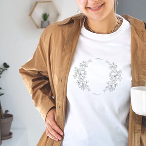 Puede incluir: Camiseta blanca con un dise&ntilde;o de corona de flores. La corona est&aacute; hecha de dibujos simples de flores y hojas.