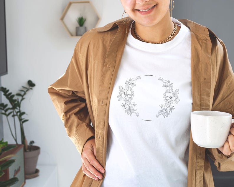 Puede incluir: Camiseta blanca con un dise&ntilde;o de corona de flores. La corona est&aacute; hecha de dibujos simples de flores y hojas.