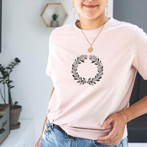 Puede incluir: Una camiseta rosa con un dise&ntilde;o de corona de flores negra.