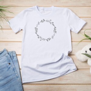 Puede incluir: Camiseta blanca con un dise&ntilde;o de corona de flores. La corona est&aacute; hecha de dibujos simples de l&iacute;neas de hojas y flores.