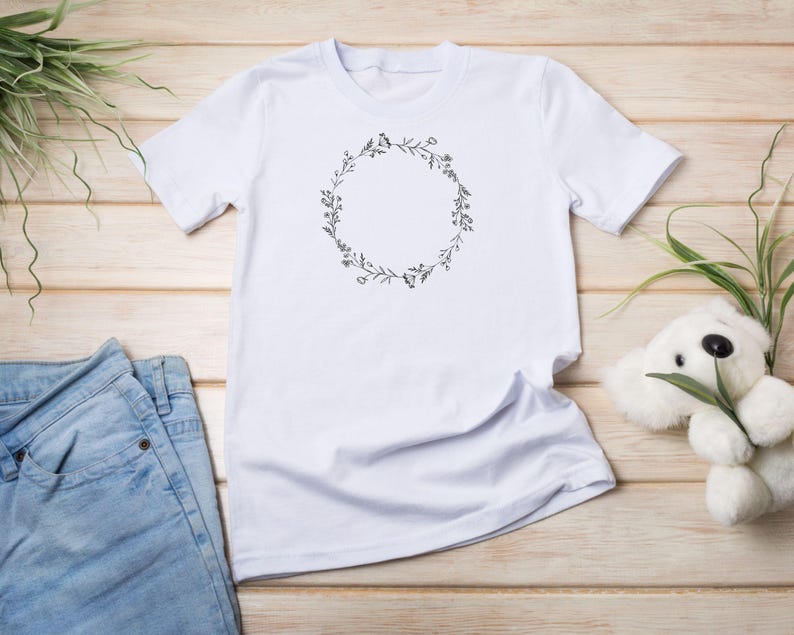 Puede incluir: Camiseta blanca con un dise&ntilde;o de corona de flores. La corona est&aacute; hecha de dibujos simples de l&iacute;neas de hojas y flores.