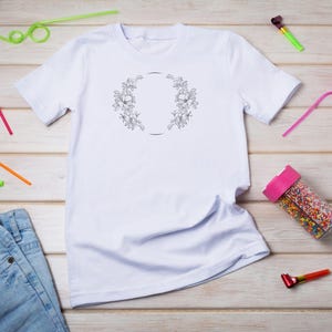 Puede incluir: Camiseta blanca con un dise&ntilde;o de corona de flores y un c&iacute;rculo en el centro. La camiseta est&aacute; colocada sobre una superficie de madera con pajitas de colores y favores de fiesta esparcidos a su alrededor.