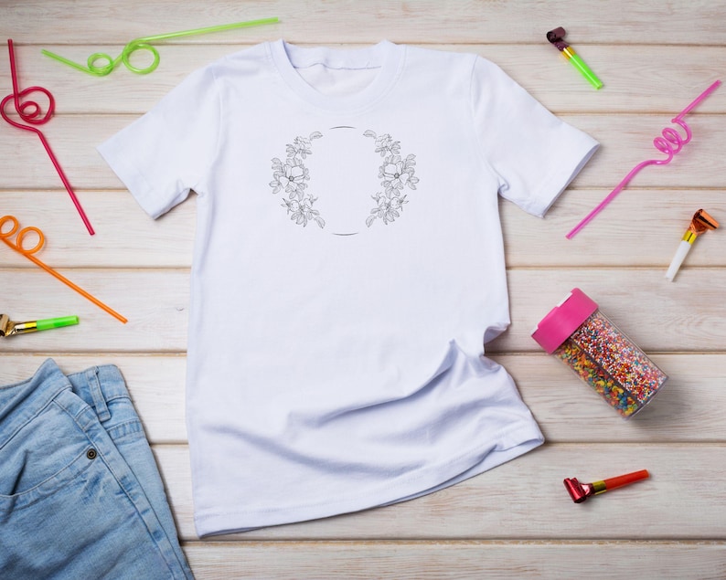 Puede incluir: Camiseta blanca con un dise&ntilde;o de corona de flores y un c&iacute;rculo en el centro. La camiseta est&aacute; colocada sobre una superficie de madera con pajitas de colores y favores de fiesta esparcidos a su alrededor.