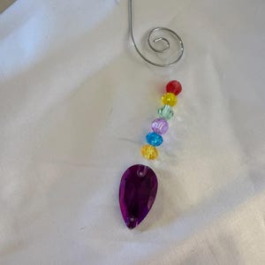 Könnte beinhalten: Dekorativer Anhänger mit einem silbernen Metallhaken und einer Kette aus bunten Perlen. Die Kette enthält rote, gelbe, grüne, blaue und violette Perlen, die zu einem großen, tropfenförmigen, facettierten lila Kristall führen. Zum Aufhängen.