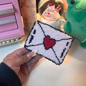 May include: White felt envelope with a red heart in the center. The envelope is outlined in black thread.
