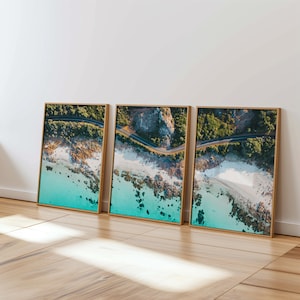 Op de afbeelding: Drie ingelijste prints met een luchtfoto van een kustweg. De afbeeldingen tonen een weg die langs een kustlijn slingert met turquoise water, wit zand en rotsachtige ontsluitingen. De frames zijn van licht hout.