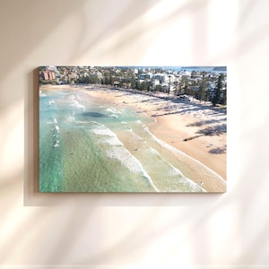 Könnte beinhalten: Eine Luftaufnahme eines Strandes mit türkisfarbenem Wasser und weißen Wellen, die an der Küste brechen. Der Strand ist von Bäumen und Gebäuden in der Ferne gesäumt.