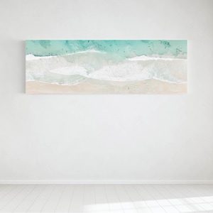 May include: A rectangular canvas print depicting a beach scene from above. The image shows turquoise water, white waves breaking on a sandy beach. Small figures of people are visible in the water and on the sand.