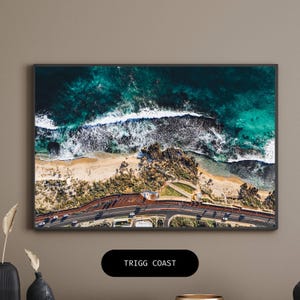 May include: An aerial view of a coastline with turquoise water crashing against a sandy beach. The waves are white and foamy, and the beach is lined with green vegetation. A road runs along the edge of the beach, with cars driving on it. The text "TRIGG COAST" is displayed below the image.