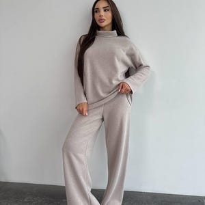 May include: A beige knitted two-piece outfit, comprising a turtleneck jumper and wide-leg trousers. The ensemble is a neutral tone, offering a relaxed style. The jumper has long sleeves, and the trousers have a loose fit.