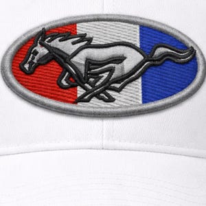 Op de afbeelding: Witte baseballpet met een geborduurde ovale patch. De patch toont een rennend paardontwerp tegen een rood, wit en blauw achtergrond, met een zilveren rand. Het paard is zilver met zwarte details.