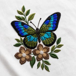 Op de afbeelding: Geborduurde vlinder met blauwe en groene vleugels, zwarte contouren en een gedetailleerd patroon. De vlinder is omgeven door bruine bloemen, groene bladeren en stelen, alles op een witte achtergrond.