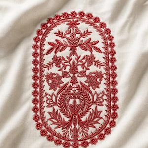 May include: An off-white fabric featuring a central oval embroidery design in deep red thread. The pattern consists of intricate floral and botanical motifs enclosed within a decorative border of small, repeating flower shapes.