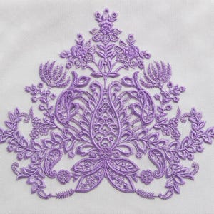 May include: A lavender-colored embroidery applique featuring a symmetrical floral and paisley design. The intricate needlework displays detailed petals, leaves, and swirling patterns, creating a decorative motif on a plain white fabric background.