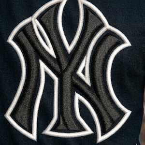 May include: Close-up of a navy blue fabric with a large, embroidered logo. The logo features the interlocking letters "NY" in black, outlined with white stitching. The design is centered and takes up a significant portion of the frame.