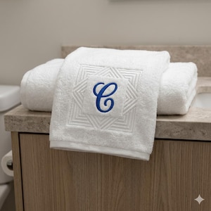 May include: A white terry cloth towel featuring a centered square embroidery pattern with a blue script letter C. The towel is folded and displays a textured fabric surface with a decorative stitched border surrounding the monogrammed letter.