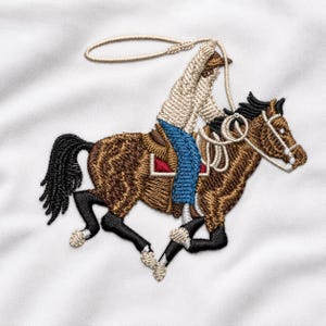 Pode incluir: Um emblema bordado apresentando um cavalo castanho com crina e cauda pretas, montado por uma figura com top bege e calças de ganga azuis. O cavaleiro segura um laço bege acima da cabeça. O cavalo tem marcas brancas nas patas inferiores.