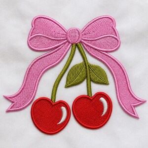 May include: An embroidered patch on white fabric featuring two bright red heart-shaped cherries with green stems and leaves, topped with a large, detailed pink ribbon bow. The cherries have white highlights, and the entire design is stitched.