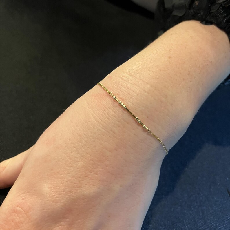 May include: A delicate gold bracelet with a linear design featuring small, evenly spaced decorative elements. The bracelet is worn on a wrist, showcasing its fine details and subtle elegance. The jewelry piece is a simple yet stylish accessory.