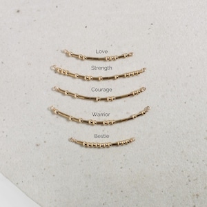 Peut inclure: Bijoux dorés avec des perles et les mots "Love", "Strength", "Courage", "Warrior" et "Bestie" écrits au-dessus de chaque pièce. Les bijoux sont disposés en forme incurvée, avec de petites perles et une barre dorée.