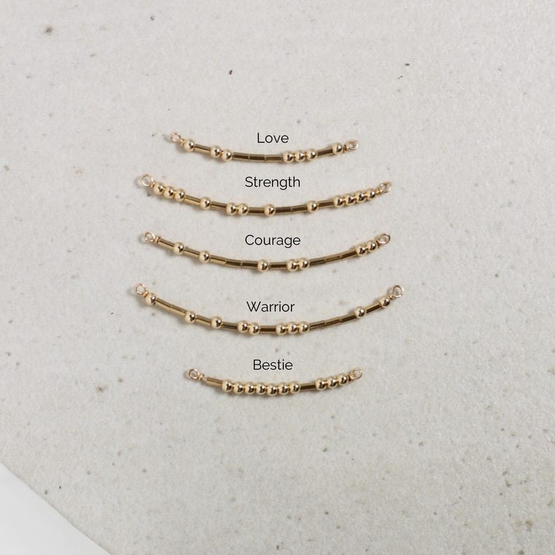 May include: Gold-colored beaded jewelry pieces with the words "Love", "Strength", "Courage", "Warrior", and "Bestie" written above each piece. The jewelry is arranged in a curved shape, with small beads and a gold bar.