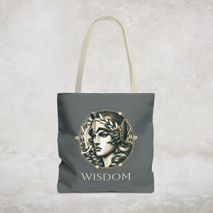 Op de afbeelding: Een grijze tote bag met een ontwerp van een vrouwengezicht in een helm en een lauwerkrans. Het ontwerp is omgeven door een gouden cirkel met een Grieks sleutelpatroon. De tekst "WISDOM" is onder het ontwerp gedrukt.