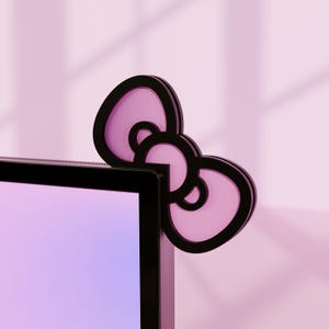 May include: A black and pink bow-shaped computer monitor decoration. The bow has a black outline with a pink interior and is placed on the corner of a black computer monitor. The background is a soft pink.