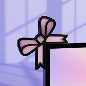 Peut inclure: Un accessoire de bureau en forme de nœud noir et rose. Le nœud est positionné sur le coin d'un moniteur noir, avec un centre rose clair et un contour noir. Le fond est un dégradé de violet et de rose.