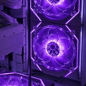 May include: A computer case interior featuring two stacked cooling fans with clear, floral-patterned frames illuminated by bright purple LED lighting. The white hardware components are visible alongside the glowing fan blades and integrated light strips.