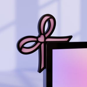 Pode incluir: Acessório decorativo em forma de laço para monitor de computador. O laço é rosa com contorno preto e um coração no centro. O acessório está preso ao canto de um monitor preto, com um fundo gradiente roxo e rosa.