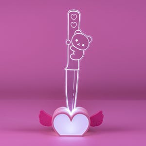 May include: A clear acrylic knife-shaped lamp featuring an etched cartoon bear holding the handle and two small hearts above it. The knife is inserted into a glowing heart-shaped base with pink wings on each side, emitting a soft white light.