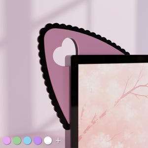 Puede incluir: Un accesorio decorativo rosa y negro para monitor con forma de orejas de gato, con un recorte en forma de corazón. El accesorio se coloca en la parte superior de un monitor negro, con una imagen rosa y blanca en la pantalla.