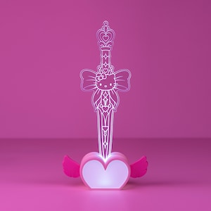 May include: A light-up acrylic sword display featuring a white line-art illustration of Hello Kitty centred on a large bow. The sword has a decorative hilt and blade, set into a pink heart-shaped base with small pink wings on each side.