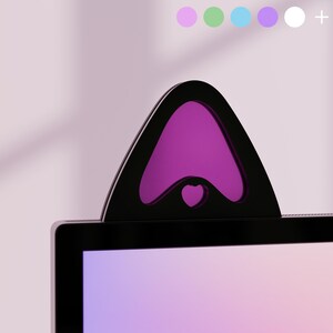Peut inclure: Accessoire d'écran noir en forme d'oreille de chat avec un intérieur violet et une découpe en forme de cœur. L'accessoire est posé sur un écran d'ordinateur. Une rangée de cercles aux couleurs pastel est visible en arrière-plan.