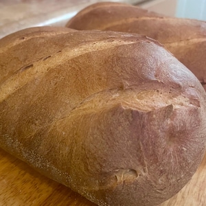 Può includere: Due pagnotte di pane appena sfornate con una crosta dorata. Le pagnotte hanno tagli diagonali sulla parte superiore e sono su una superficie di legno. Il pane sembra essere in stile rustico, ideale per panini o toast.
