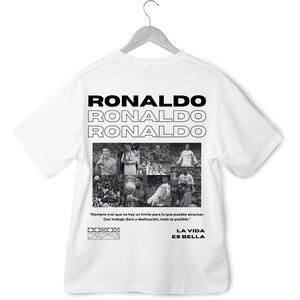 May include: White t-shirt with a black and white photo collage of a soccer player. The text "RONALDO" is repeated three times in a stacked format. The quote "Siempre crei que no hay un limite para lo que puedes alcanzar. Con trabajo duro y dedicaci&oacute;n, todo es posible." is printed below the collage. The text "LA VIDA ES BELLA" is printed below the quote.