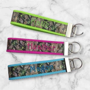 May include: Three personalized keychains with camouflage fabric and a silver key ring. The keychains are green, pink, and blue with the names "Katie", "Heather", and "Magen" embroidered in white.
