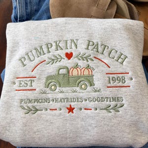 Puede incluir: Una sudadera gris claro con bordados en verde y naranja que dicen PUMPKIN PATCH EST 1998, un camión verde que lleva dos calabazas, un corazón rojo y el texto PUMPKINS HAYRIDES GOODTIMES con una pequeña estrella naranja de adorno.