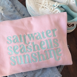 May include: A light pink sweatshirt with the embroidered words "saltwater seashells sunshine" in light blue. The sweatshirt is folded on top of a pair of light blue denim shorts. White athletic shoes with light blue accents are visible in the background.