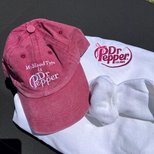 May include: A red baseball cap with the text "My Blood Type is Dr Pepper" embroidered on the front. A white t-shirt with a pink and red Dr Pepper logo embroidered on the front. The logo includes the text "Dr Pepper Est. 1885".