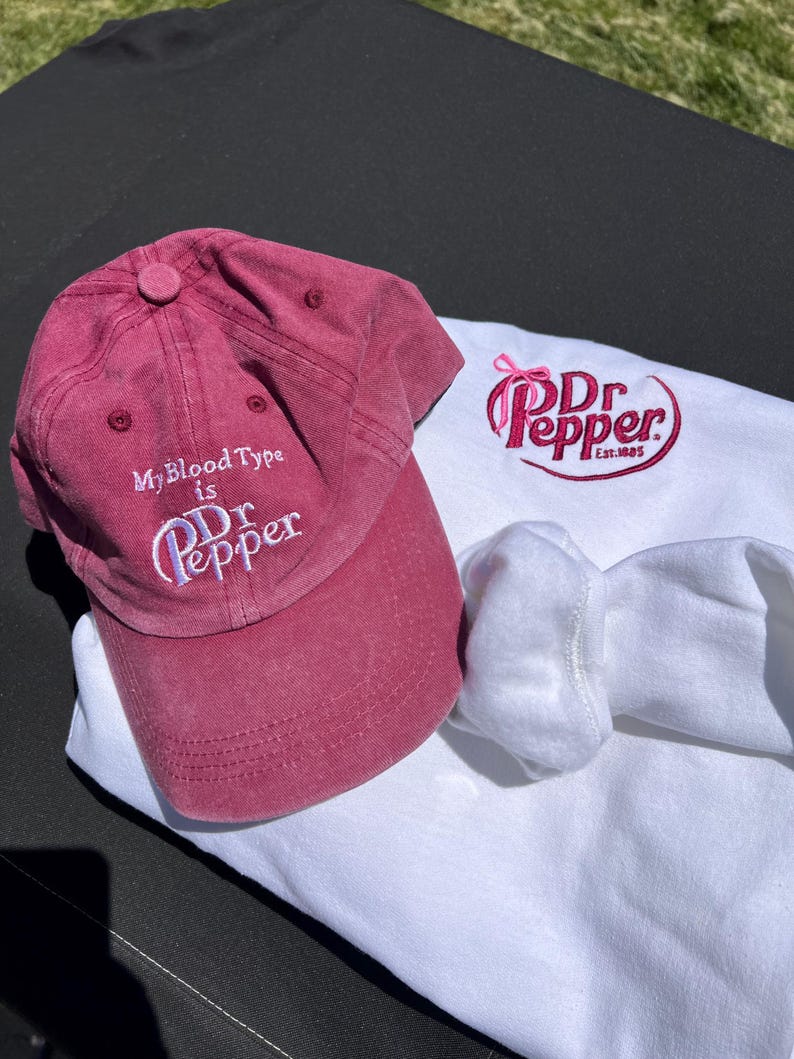 May include: A red baseball cap with the text "My Blood Type is Dr Pepper" embroidered on the front. A white t-shirt with a pink and red Dr Pepper logo embroidered on the front. The logo includes the text "Dr Pepper Est. 1885".