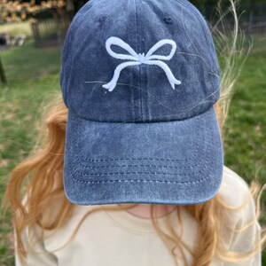 May include: A dark blue denim baseball cap with a white embroidered bow on the front.