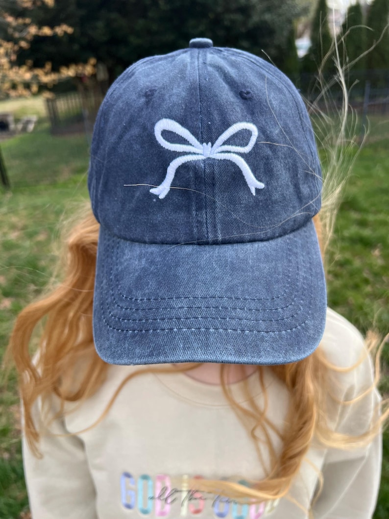 May include: A dark blue denim baseball cap with a white embroidered bow on the front.
