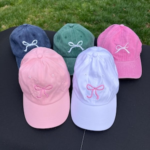May include: Five baseball caps in different colors with a white bow embroidered on each. The caps are arranged in a semi-circle on a black surface.