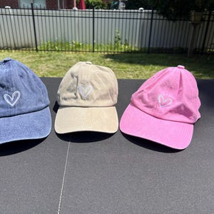 May include: Three baseball caps with a white heart design. One cap is blue, one is beige, and one is pink.