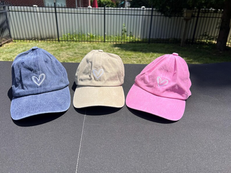 May include: Three baseball caps with a white heart design. One cap is blue, one is beige, and one is pink.