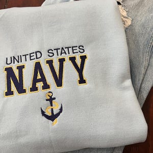 May include: A light blue sweatshirt with the words "UNITED STATES NAVY" embroidered in navy blue and gold. An anchor emblem is embroidered below the text. The sweatshirt is folded next to a pair of light blue denim jeans.