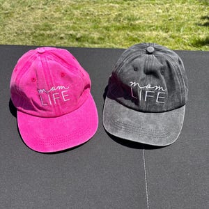 May include: Two baseball caps, one pink and one black, with the text "mom life" embroidered in white on the front.