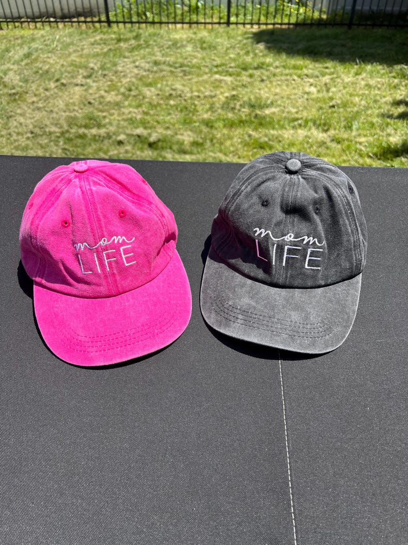 May include: Two baseball caps, one pink and one black, with the text "mom life" embroidered in white on the front.