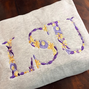 May include: A light gray sweatshirt featuring the letters LSU embroidered in a floral pattern. The letters are formed by yellow and purple flowers, leaves, and vines, creating a decorative botanical design across the front of the garment.
